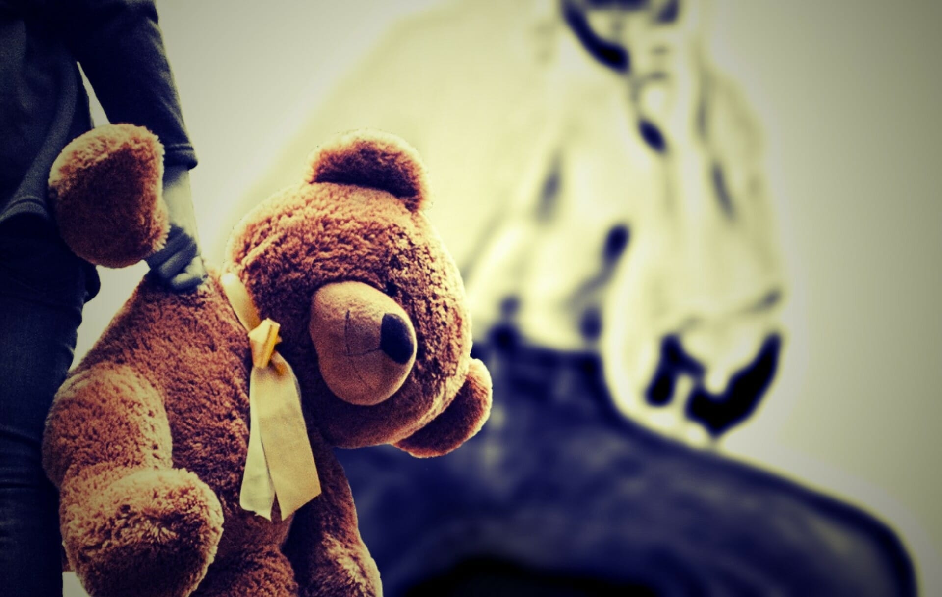 Kind mit Teddybären in der Hand. Im Hintergrund sieht man die Umrisse einer männlichen Person. Das Kind hat Angst vor Gewalt und Missbrauch.